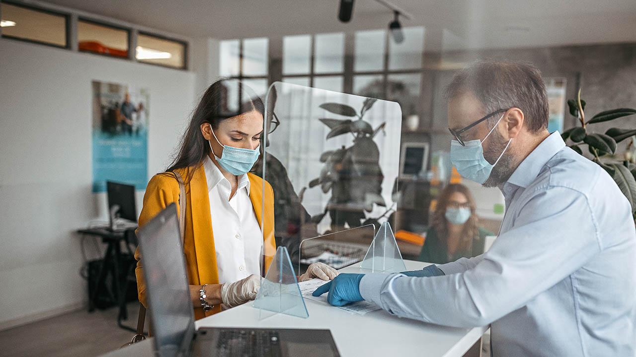 Bank teller discussing paperwork with customer at bank counter wearing protective gloves and face mask. Office with acrylic glass partition on desk. Acrylic glass wall - protection against coughs and spitting, protection against viruses.