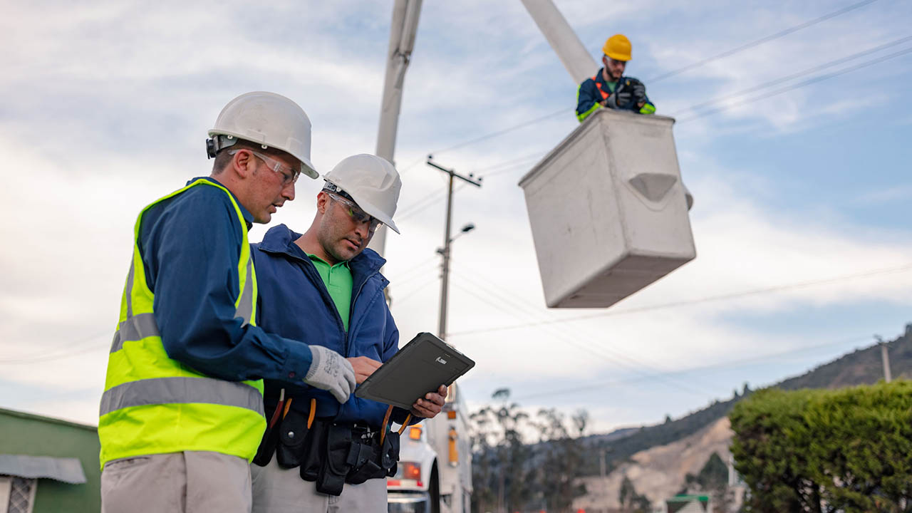 Two utility workers look at a tablet on the ground while a lineman looks at a handheld mobile computer from the bucket above